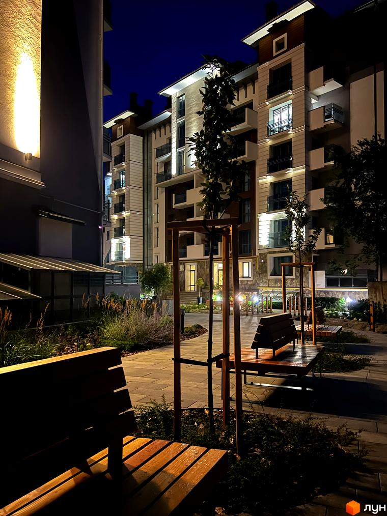 Вечірній вигляд на багатоповерхові будинки Комплексу Крайобраз з освітленими фасадами та балконами, а також облаштованою прибудинковою територією з лавками.