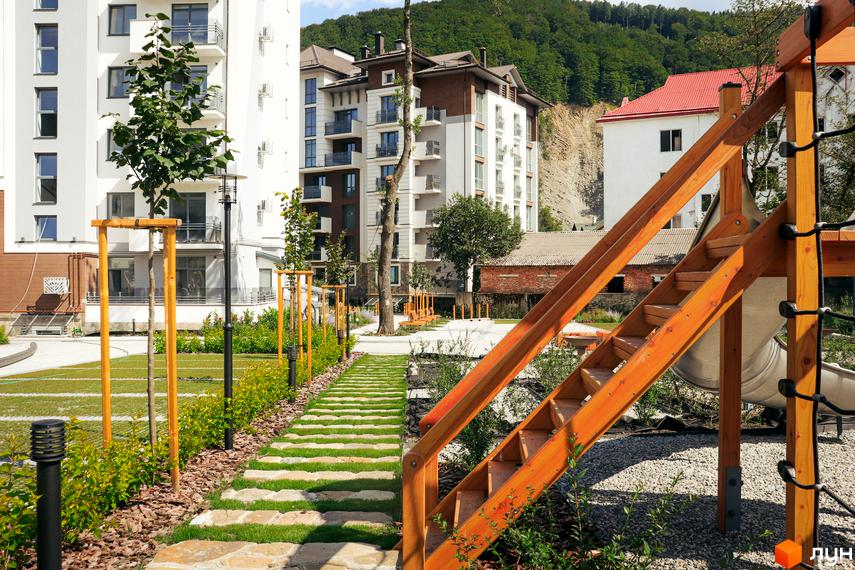 Фасади багатоповерхових будинків Комплексу Крайобраз у білих та коричневих тонах. Прибудинкова територія з дитячим майданчиком та стежкою з каменів серед зелені.