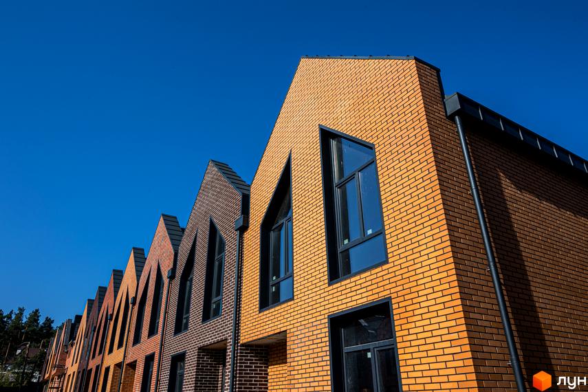 Сучасні таунхауси FORT HOMES з цегляним фасадом та незвичайними вікнами підкреслюють унікальну архітектуру комплексу. Будинки розташовані вздовж вулиці під яскравим синім небом.