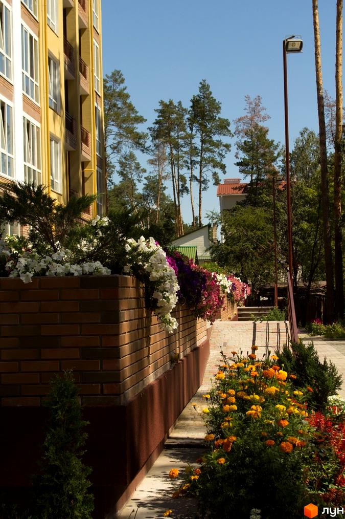 Житловий комплекс Женев'єва: жовта багатоповерхівка з білими вікнами та балконами, прибудинкова територія з квітковими клумбами та деревами.
