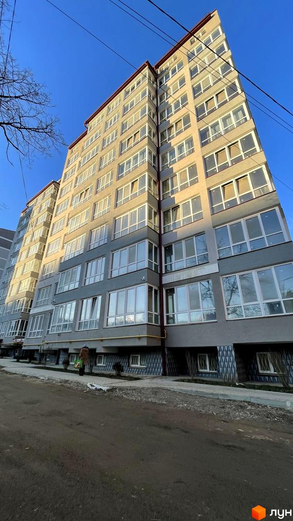 Високий житловий будинок з багатьма вікнами та світлим фасадом, розташований біля ґрунтової дороги та тротуару.