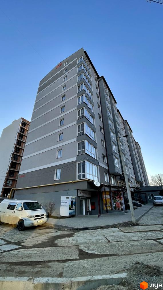 Зовнішній вигляд новобудови на вул. Шухевича, 3б, з сірими фасадами та скляними балконами. Поруч прибудинкова територія з паркуванням.