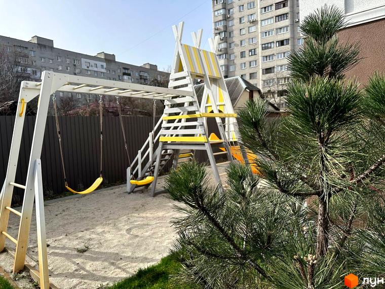 Житловий комплекс SMART IDEA з дитячим майданчиком та гойдалками на піщаному покритті. На задньому плані видно багатоповерхові житлові будинки.