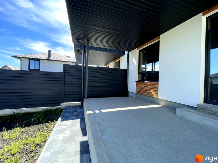 Фасад Житлового комплексу Woodland Hill: білі стіни, чорні віконні рами, цегляні вставки. Поруч бетонний майданчик, доріжка з бруківки та темний паркан.