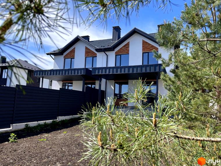 Зображення Дуплекси Woodland Hill: білий дуплекс з темним дахом, цегляними вставками та навісом. На передньому плані сосни та темний паркан.