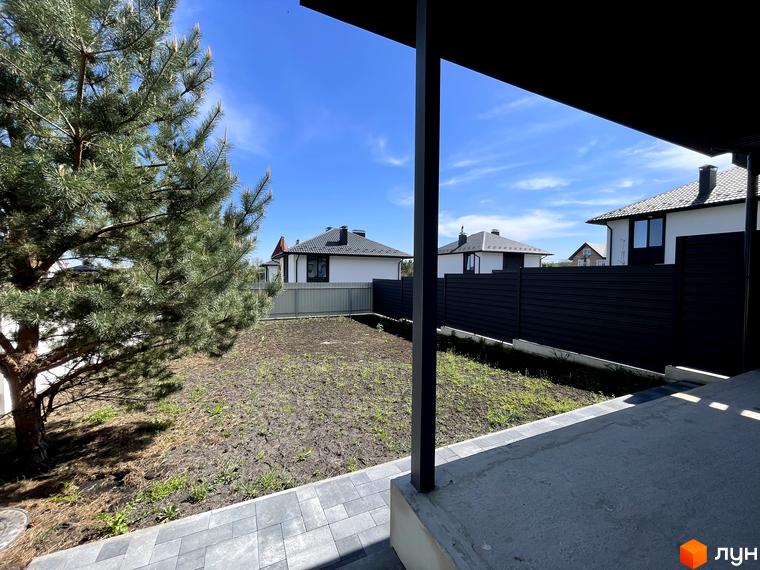 Дуплекси Woodland Hill з білими фасадами та темними дахами, оточені чорним парканом. На ділянці видно ґрунт, бруківку та велике хвойне дерево.