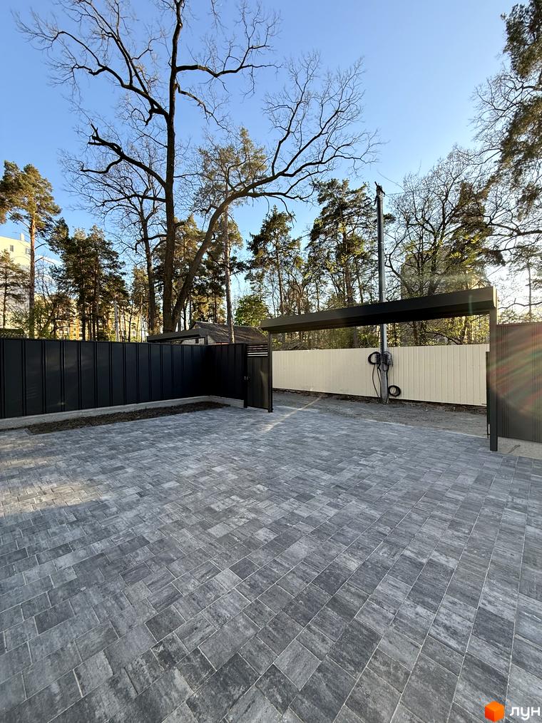 Зовнішній вигляд новобудови КМ М-8А з бруківкою та навісом для авто. Територія комплексу з парканом та деревами.