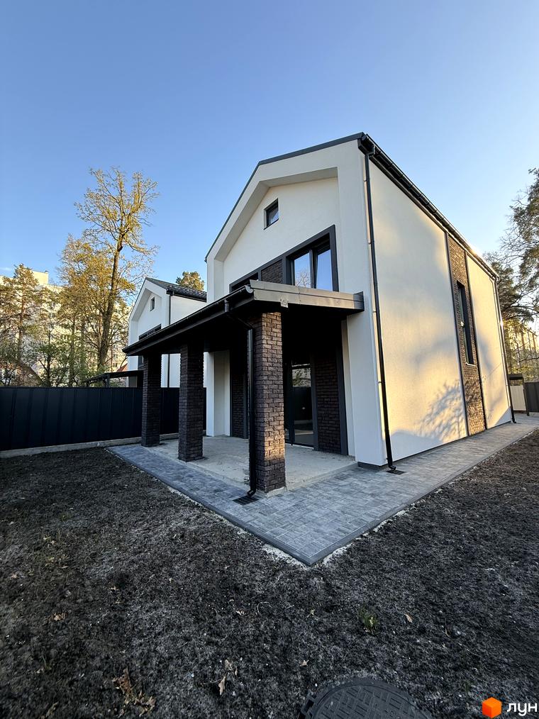 Котеджне містечко КМ М-8А: двоповерховий будинок з білою штукатуркою, темною цегляною обробкою та навісом на цегляних колонах. Прибудинкова територія з бруківкою та газоном.