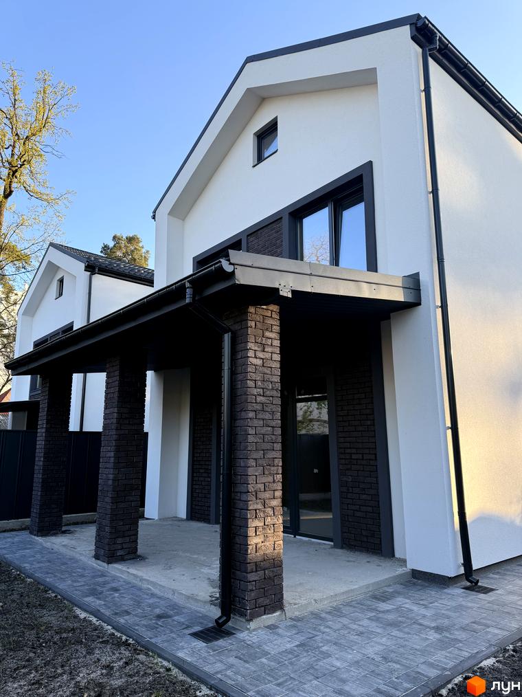 Котеджне містечко КМ М-8А: фасад будинку з білою штукатуркою, темною цеглою та чорною покрівлею. Навколо будинку викладена бруківка та зелена ділянка.