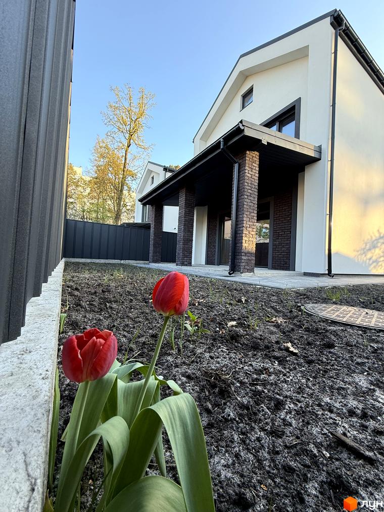 Котеджне містечко КМ М-8А: білий двоповерховий будинок з цегляними колонами та червоними тюльпанами на тлі блакитного неба.