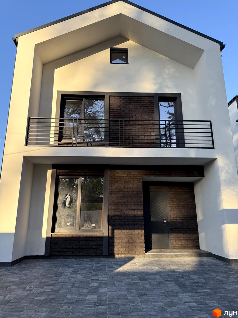 Двоповерховий будинок Котеджного містечка М-8А зі світлими стінами, темним дахом, коричневою цегляною обробкою та балконом. Перед будинком укладена сіра тротуарна плитка.