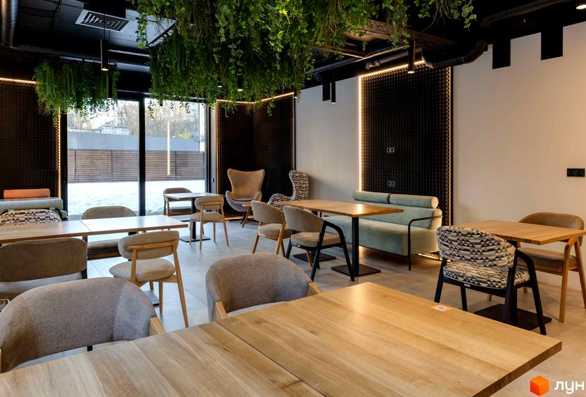 Інтер'єр кафе з дерев'яними столами та кріслами. Зверху звисають зелені рослини, створюючи затишну атмосферу в приміщенні.