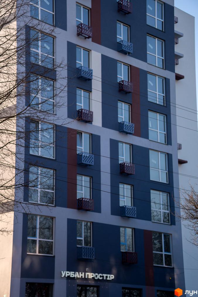 Новобудова ЗиґЗаґ з синьо-сірим фасадом, великими вікнами та декоративними кошиками для кондиціонерів. Поруч розташовані дерева та електричні дроти.