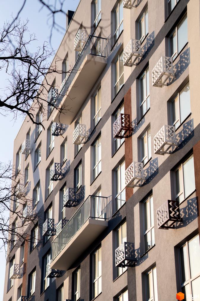 Новобудова ЗиґЗаґ з сірим фасадом, балконами та декоративними коробами для кондиціонерів. На передньому плані видно гілки дерев.