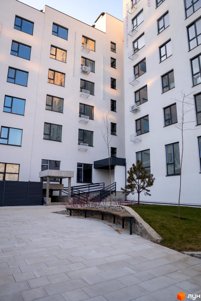 Зовнішній вигляд новобудови ЖК Синергія Light з білими фасадами та великою кількістю вікон. Прибудинкова територія з вимощеним майданчиком, лавкою та зеленими насадженнями.