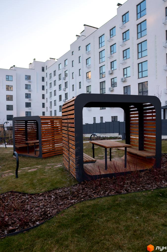 Зовнішній вигляд білосніжних багатоповерхових будинків ЖК Синергія Light з численними вікнами та кондиціонерами. На прибудинковій території розташовані дерев'яні альтанки з лавками та столами.