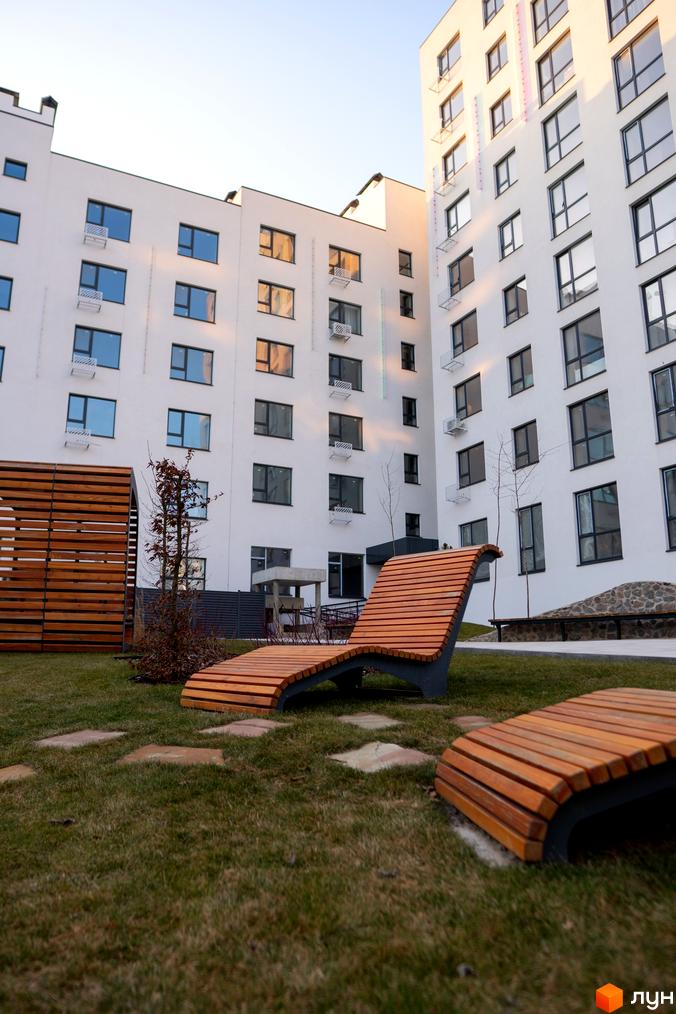 Новобудова Синергія Light з білим фасадом та великими вікнами. На прибудинковій території зелена галявина з дерев'яними лежаками та доріжками з плитки.