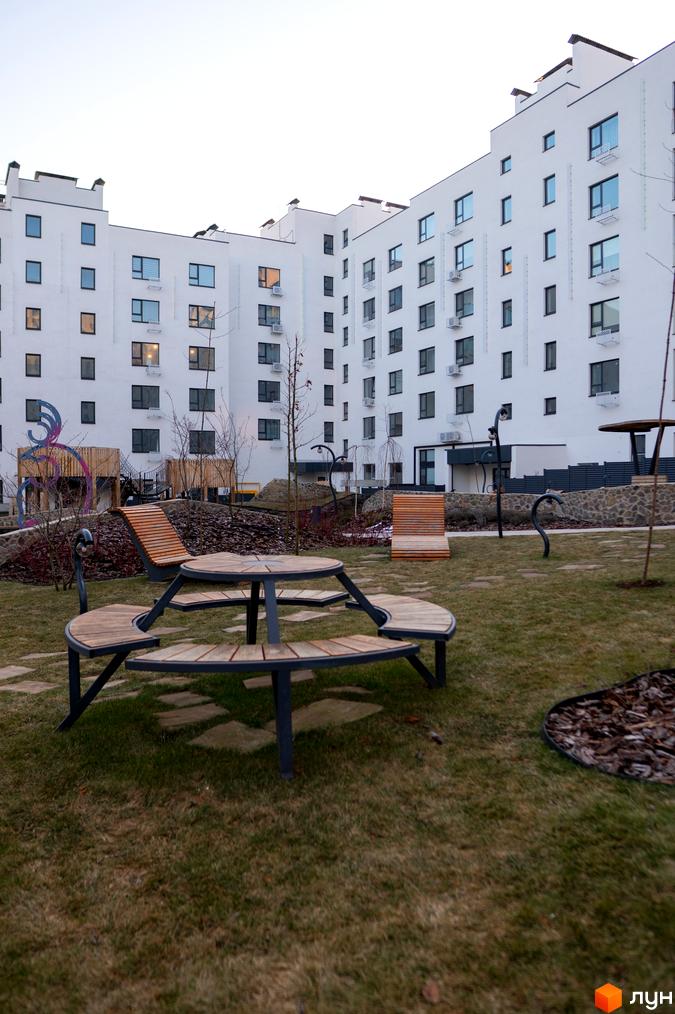 Житловий комплекс Синергія Light з білими фасадами та кондиціонерами. Прибудинкова територія з дерев'яними лавками та столом, газоном та декоративними елементами.