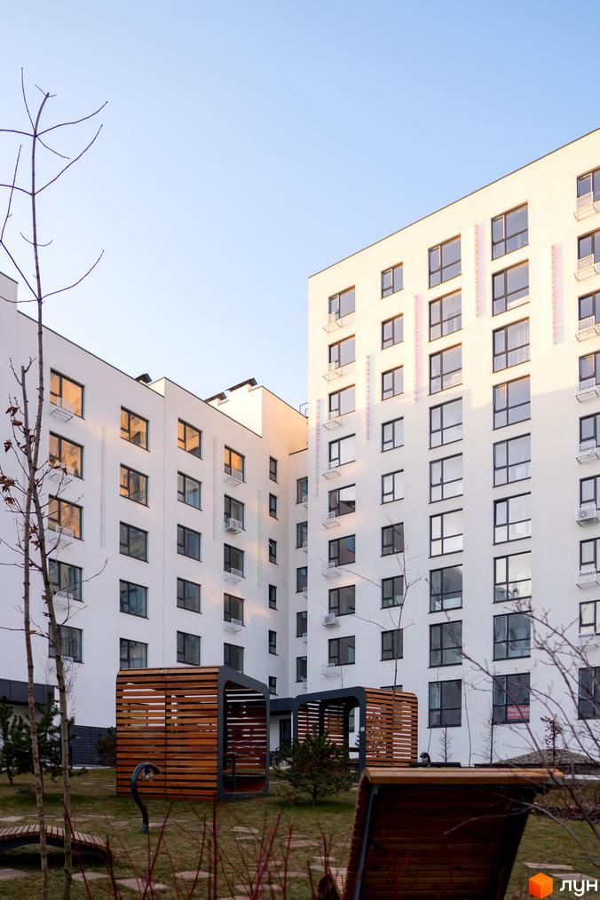 Зовнішній вигляд новобудови ЖК Синергія Light з білими фасадами та великими вікнами. На прибудинковій території розташовані дерев'яні альтанки та лавки.