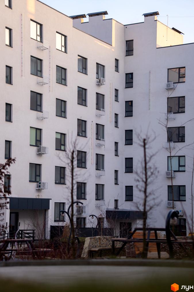 Фасад житлового комплексу Синергія Light з численними вікнами та кондиціонерами. Прибудинкова територія з лавками та деревами.