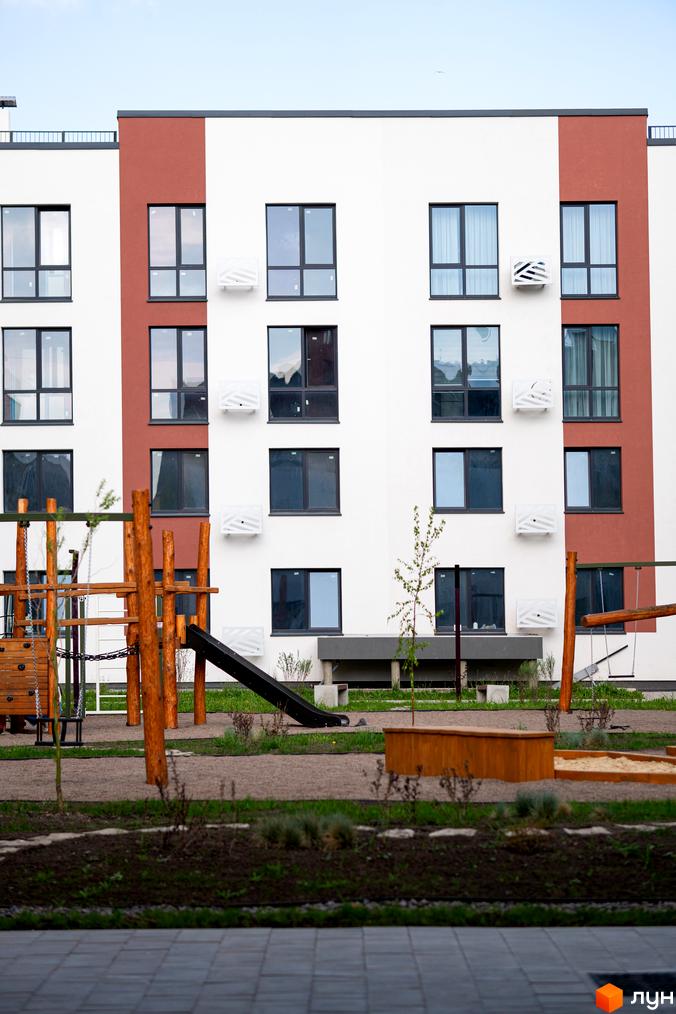 Зовнішній вигляд новобудови ЖК Субурбія з вікнами та кондиціонерами, поруч дитячий майданчик з гіркою та гойдалкою.