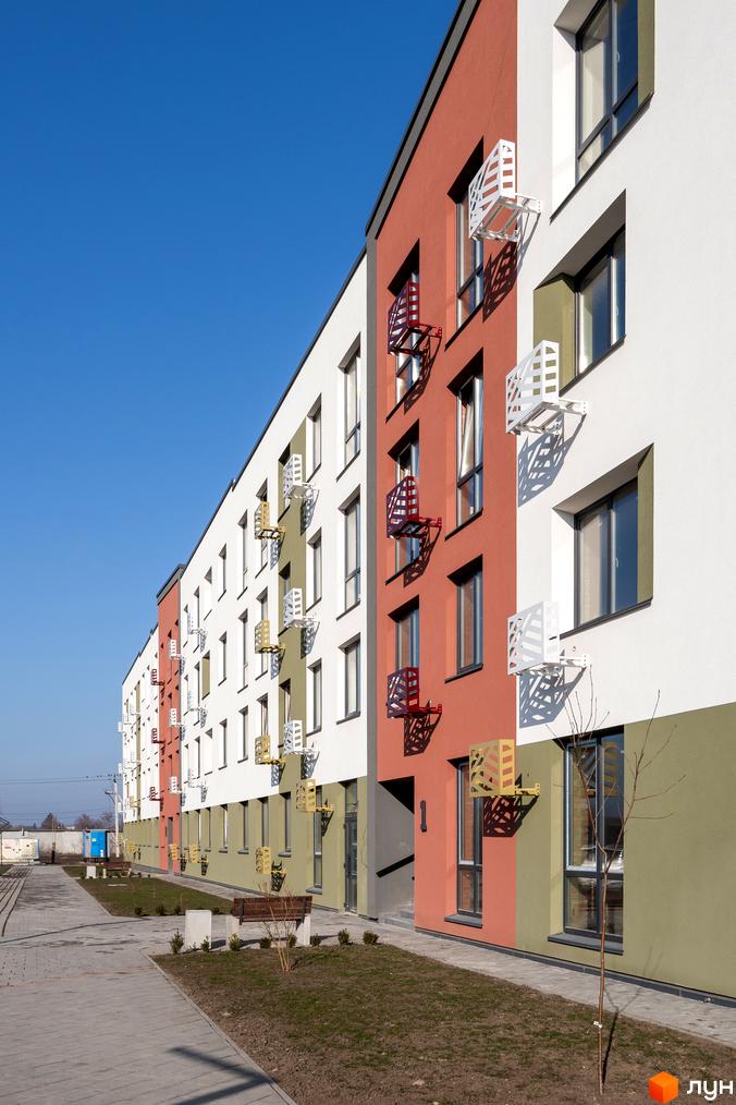 Зовнішній вигляд будинку житлового комплексу Субурбія з білими та червоними фасадами, вікнами та декоративними елементами. Поруч розташована доглянута прибудинкова територія з доріжками та лавками.