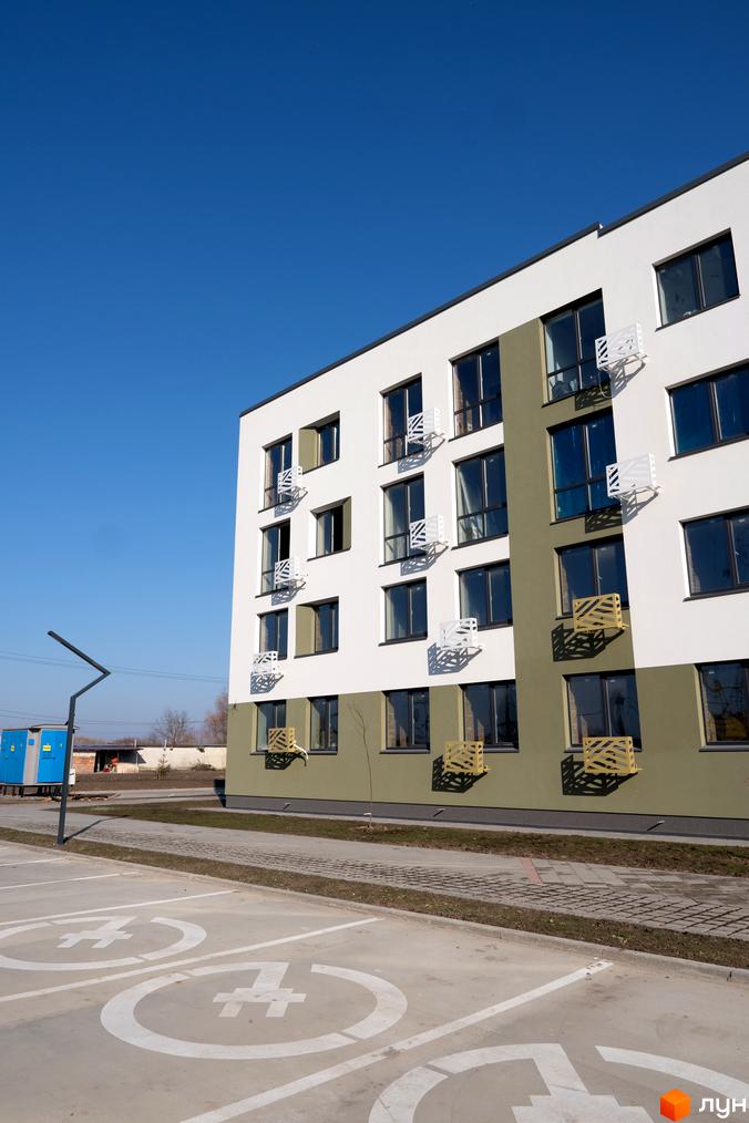 Будинок житлового комплексу Субурбія з білими та зеленими фасадами, вікнами та кондиціонерами, на тлі блакитного неба. Поруч парковка з розміткою.