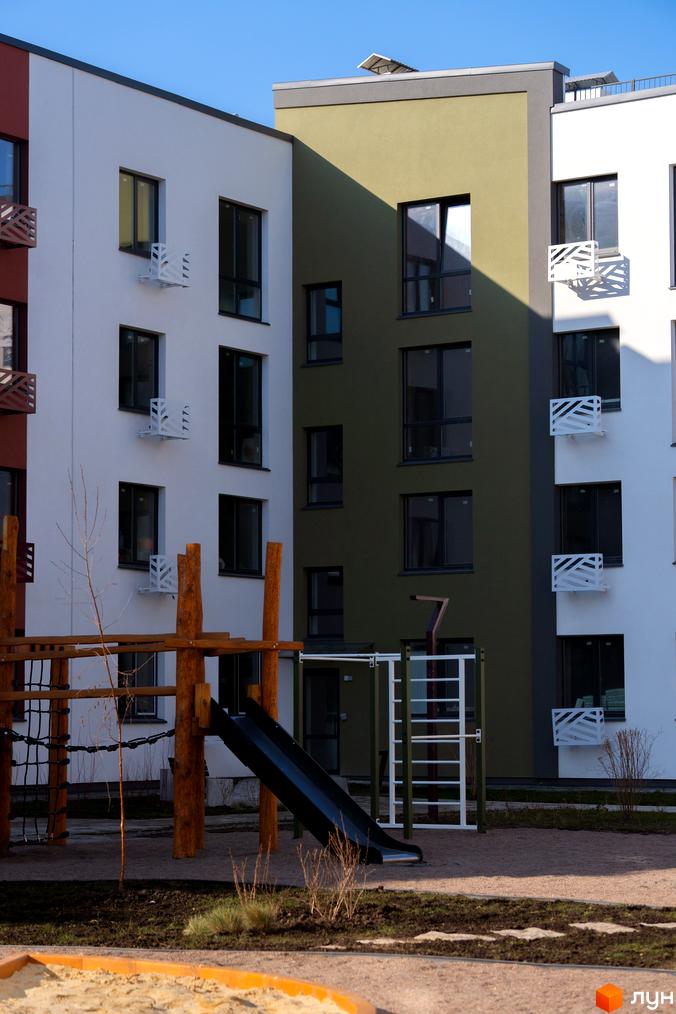Зовнішній вигляд новобудови ЖК Субурбія з білими та оливковими фасадами, вікнами та балконами. Прибудинкова територія з дитячим майданчиком, гіркою та пісочницею.