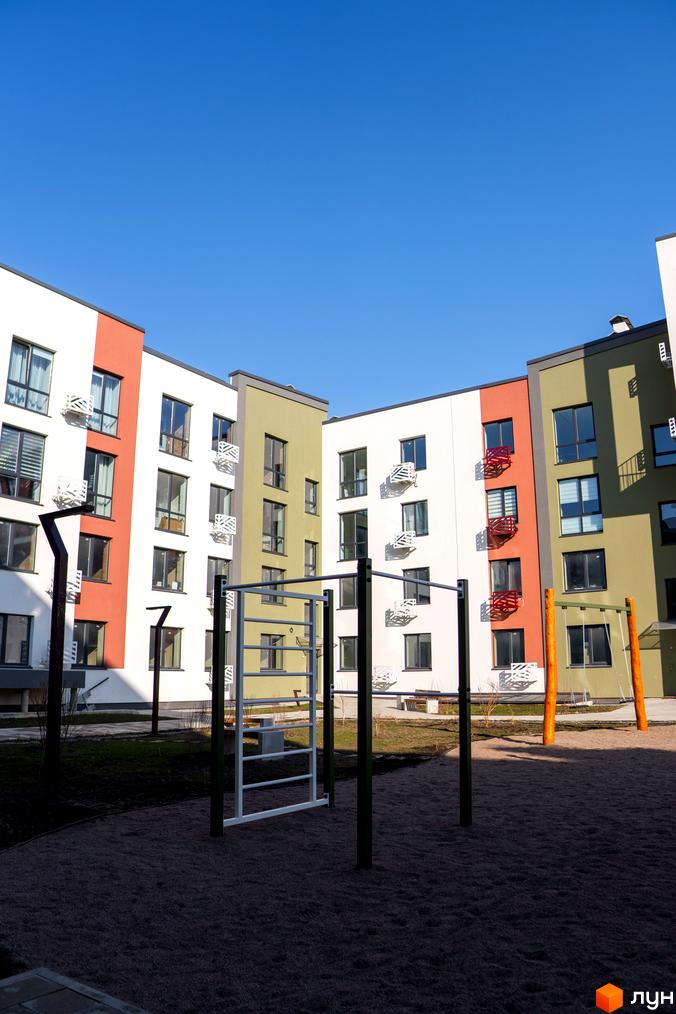 Житловий комплекс Субурбія з фасадами білого, червоного та оливкового кольорів. На прибудинковій території розташовані спортивні снаряди та зелені насадження.