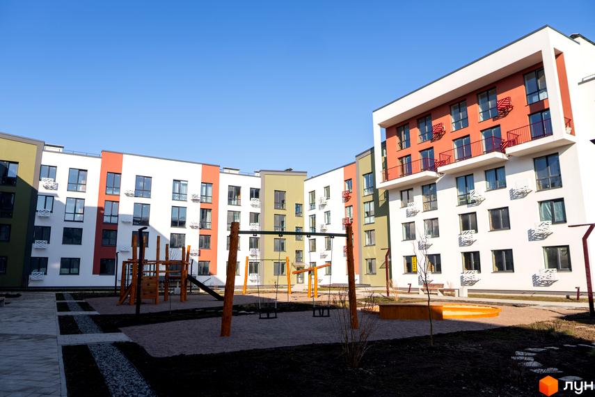 Житловий комплекс Субурбія з фасадами білого, помаранчевого та зеленого кольорів. На прибудинковій території розташований дитячий майданчик з гойдалками та пісочницею.