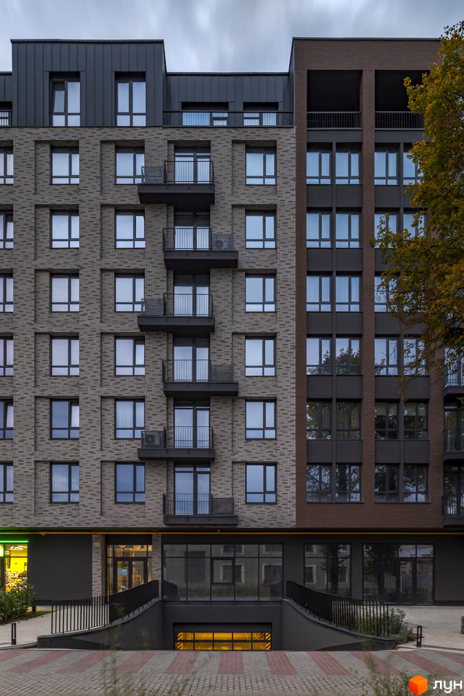 Фасад житлового комплексу Avalon Yard, оздоблений цеглою та темними панелями, з балконами та великими вікнами. Навколо будинку розташована бруківка та дерева.