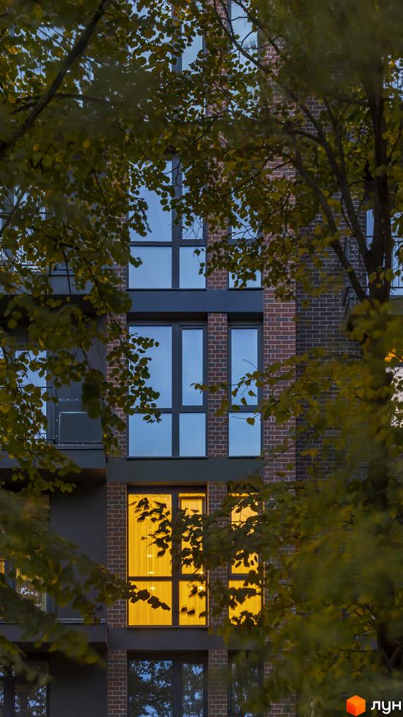 Фасад житлового комплексу Avalon Yard з цегляним оздобленням та великими вікнами, крізь які видно жовте світло. Навколо будівлі розташовані дерева з осіннім листям.