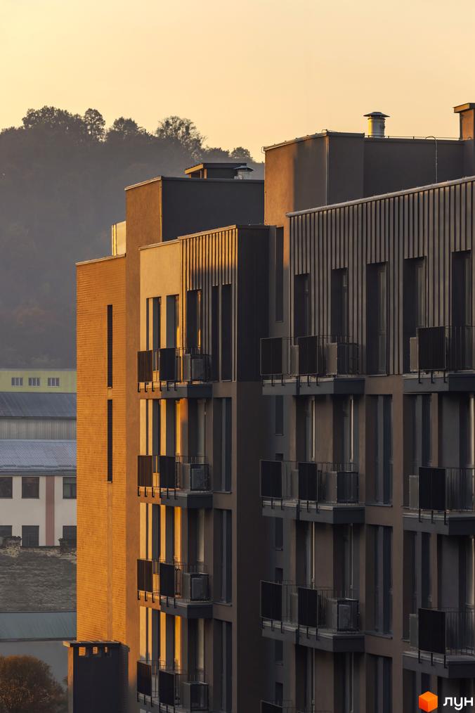 Фасад новобудови ЖК Avalon Yard з цегляним оздобленням та балконами, освітлений сонцем на заході. Вид на будівлі та схил гори.