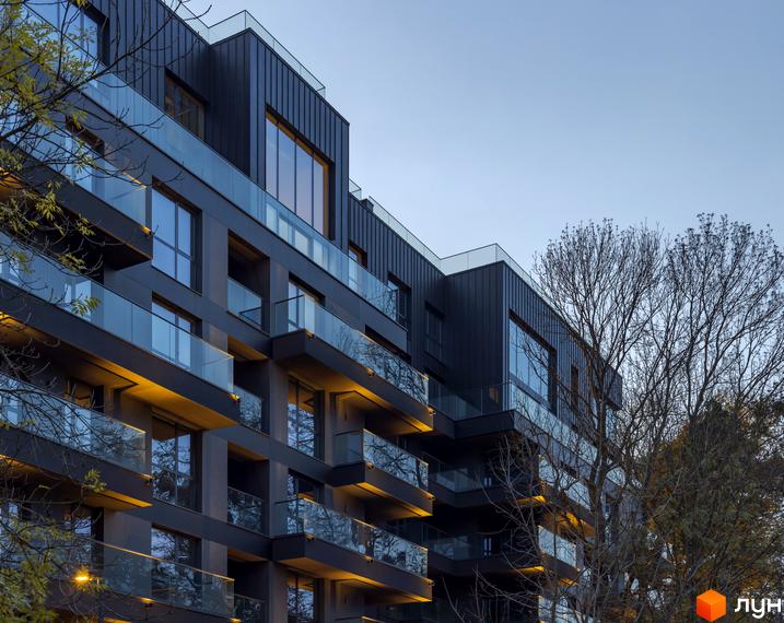 Фасад житлового комплексу Avalon Prime з скляними балконами та темними панелями. Навколо будівлі ростуть дерева, що додають природного колориту.