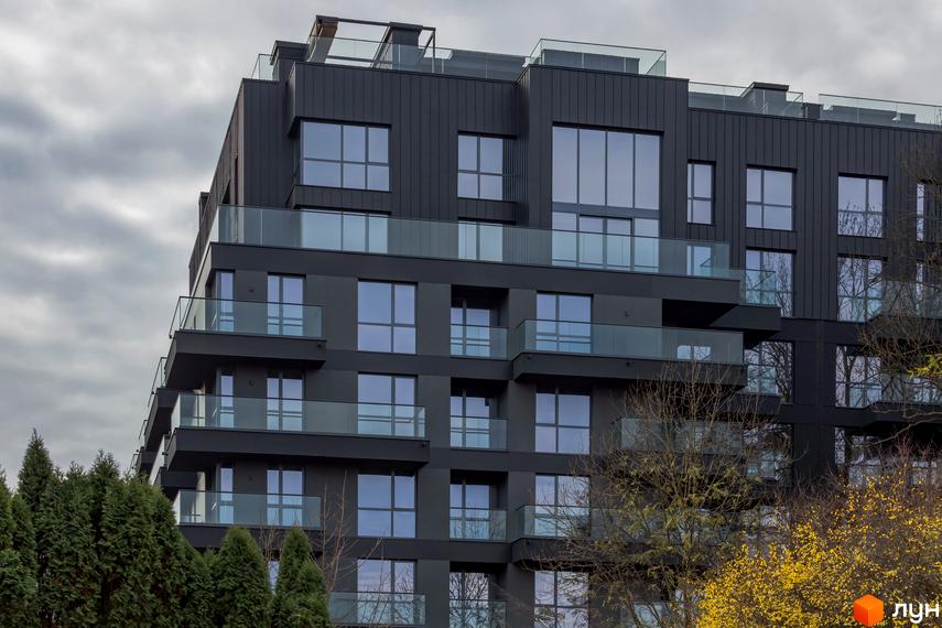 Зовнішній вигляд ЖК Avalon Prime з темним фасадом та скляними балконами. Навколо будівлі зелені дерева та хмарне небо.