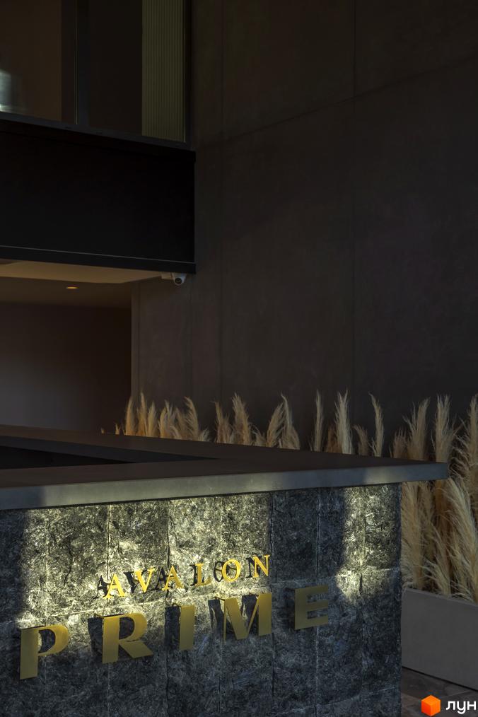 Житловий комплекс Avalon Prime з золотими літерами на фасаді з сірого каменю. Поруч видніються декоративні рослини та вхідна група.