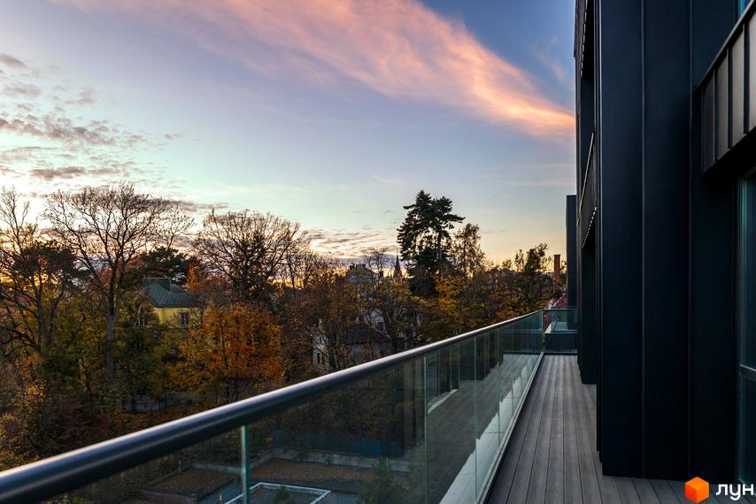 Вид з балкона на ЖК Avalon Prime з видом на дерева восени та хмари на заході сонця. Сучасна архітектура будинку з темними фасадами та скляними перилами.