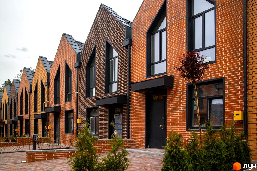 Таунхауси FORT HOMES з цегляним фасадом та трикутними вікнами, розташовані на вимощеній бруківкою території з зеленими насадженнями.