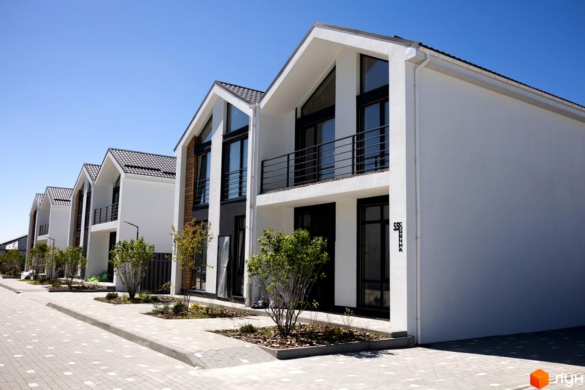 Зовнішній вигляд котеджів КМ Gardd House Eco Village з білими фасадами та чорними вікнами, розташованих вздовж вимощеної вулиці з зеленими насадженнями.