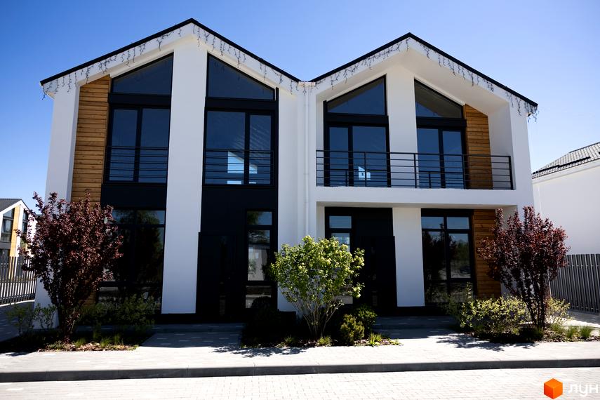 Двоповерховий будинок білого кольору з чорними вікнами та дерев'яними вставками. Перед ним бруківка та зелені насадження КМ Gardd House Eco Village.