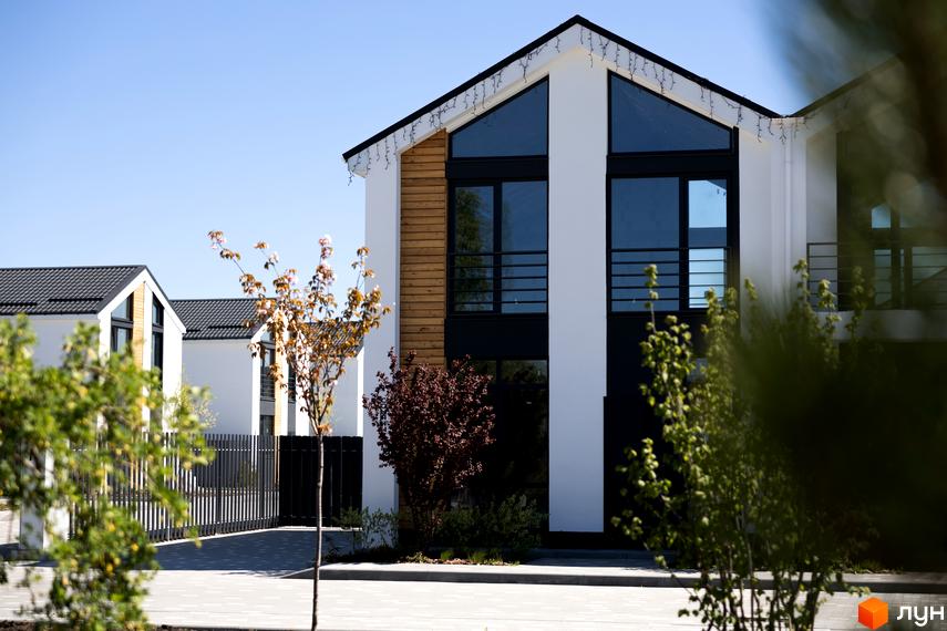 Будинки Котеджного містечка Gardd House Eco Village з білими фасадами, чорними дахами, великими вікнами та дерев'яними вставками, оточені бруківкою та деревами.