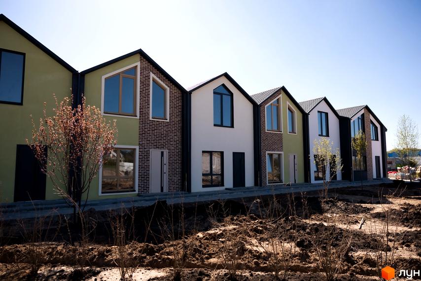 Ряд будинків з двосхилими дахами та великими вікнами. Перед будинками земляні роботи та рослини, що висаджуються.