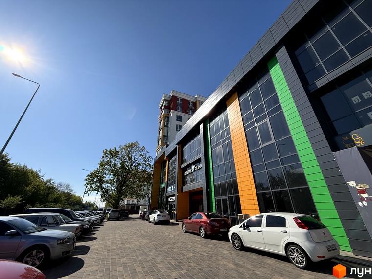 Зовнішній вигляд будівлі Житлового комплексу Добре Місто 2 з великими вікнами та кольоровими вставками. Перед будинком парковка з автомобілями.