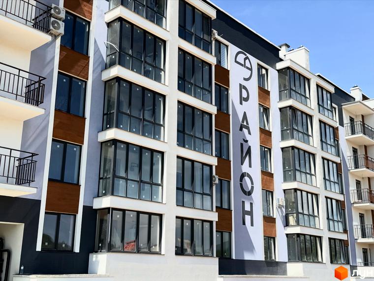 Новобудова ЖК 9 Район має біло-сірий фасад з коричневими вставками та великими панорамними вікнами. Будинок розташований на фоні блакитного неба.