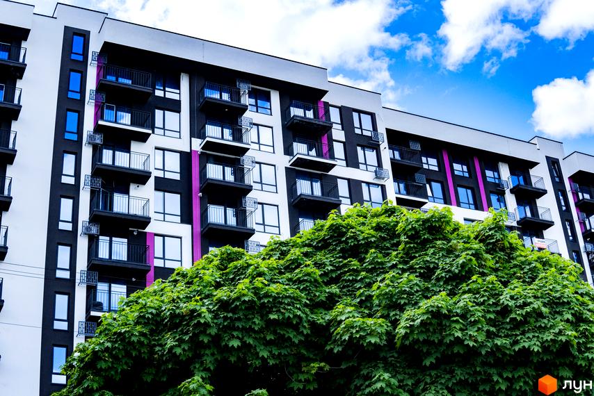 Зовнішній вигляд Новобудови IQ House з балконами та великими вікнами на фоні блакитного неба. Перед будинком зелені дерева.