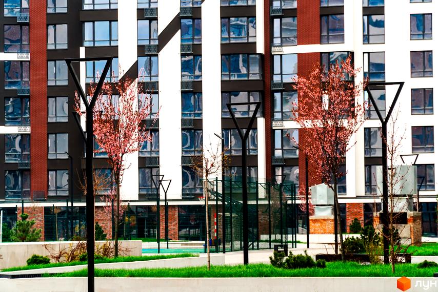 Житловий район Manhattan: фасади будівель з цегляними та білими вставками, великою кількістю вікон. Прибудинкова територія з деревами, що квітнуть, газоном та спортивним майданчиком.