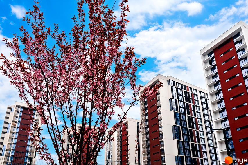 Житловий район Manhattan: сучасні новобудови з червоними та білими фасадами на тлі блакитного неба з хмарами та квітучим деревом.