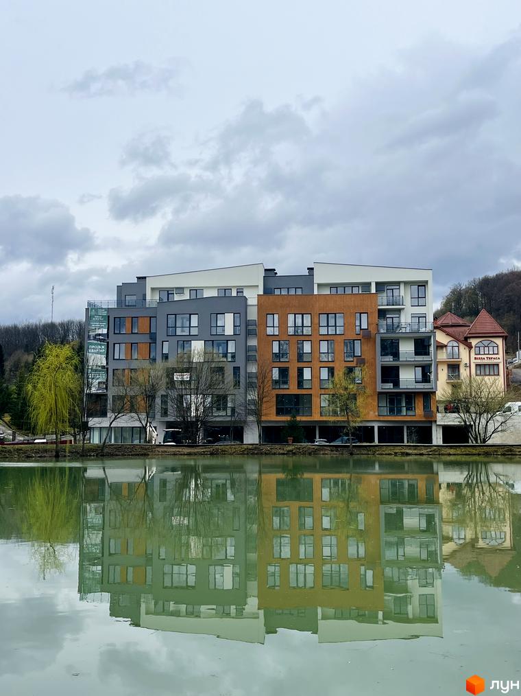 Житловий комплекс Forest Residence Polyana на березі озера з відображенням у воді. Будинки з різнокольоровими фасадами та великими вікнами, поруч дерева та зелень.