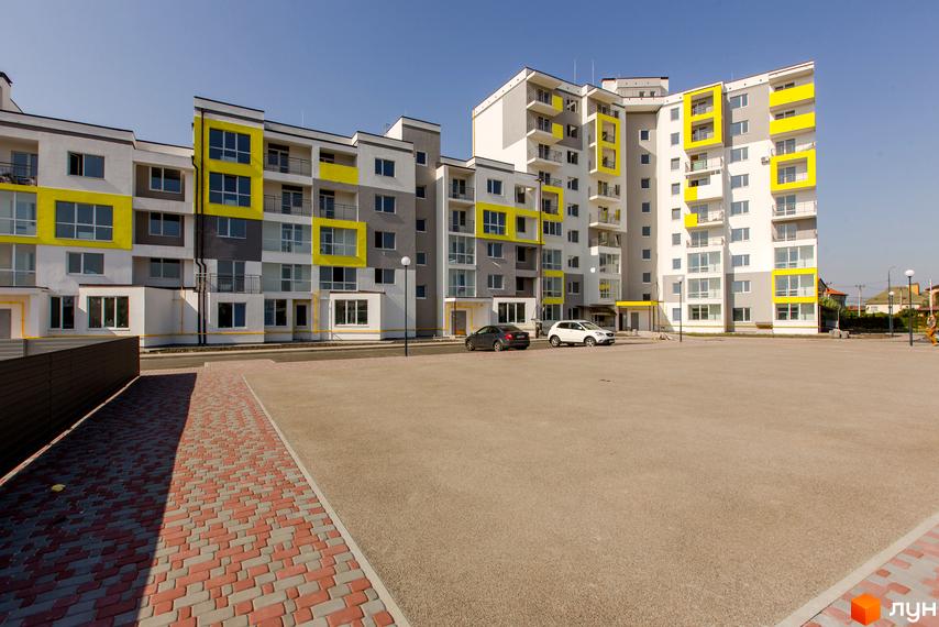 Житловий комплекс Сан Сіті з яскравими жовтими акцентами на фасаді. Перед будинком розташований просторий паркінг та доріжка з бруківки.