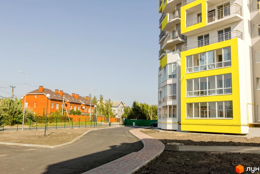 Житловий комплекс Сан Сіті. Зовнішній вигляд новобудови з жовтими елементами та балконами, поруч розташовані цегляні будинки та доглянута прибудинкова територія з доріжками.